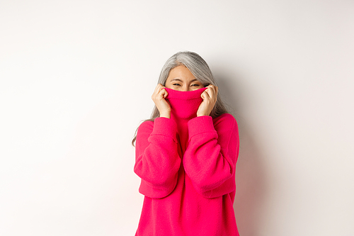 Cheerful asian senior woman laughing, wearing warm sweater, hiding face inside collar, standing happy over white background.