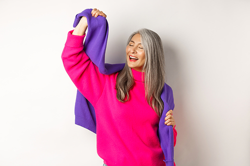 Carefree korean elderly woman in pink sweater, dancing with sweatshirt on shoulders and smiling, posing happy over white background.