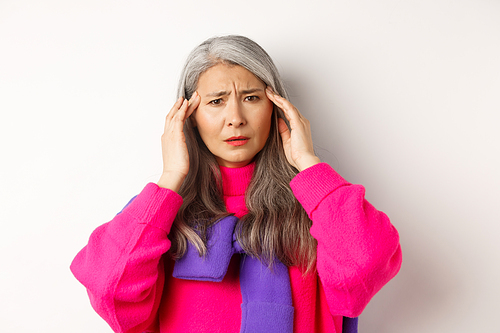 Close-up of fashionable asian senior woman feeling unwell, touching head and grimacing from headache, complain on migraine, white background.