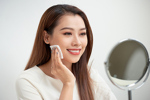 Taking off her make-up. Beautiful cheerful young woman using cotton disk and looking at her reflection in mirror with smile while sitting at the dressing table