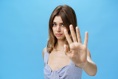 Silly and shy insecure attractive girl pulling hand towards to cover face from camera making moody timid expression asking to stop not liking what happening saying no with body language over blue wall. Lifestyle.