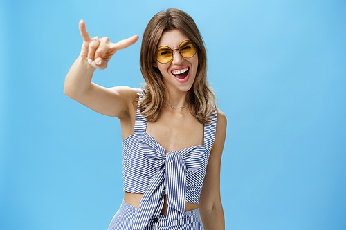 Waist-up shot of confident and daring charming young woman in matching trendy clothes yelling yeah showing rock-n-roll gesture and gazing at camera wearing cool sunglasses enjoying party or festival.Lifestyle.