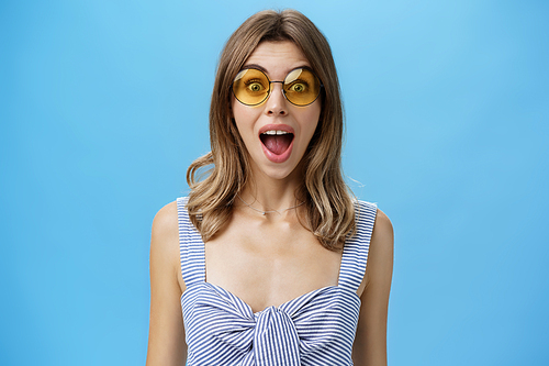 Waist-up shot of excited surprised and emotive charismatic caucasian woman in trendy sunglasses opening mouth from amazement and joy gazing at camera impressed and astonished over blue background. Lifestyle.