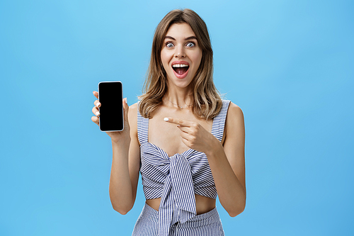 Excited happy woman with gapped teeth finally bought brand new smartphone holding device in hand pointing at cellphone screen showing cool app smiling broadly from joy against blue wall. Advertisement concept