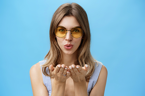Close-up shot of feminine and tender charming woman in trendy yellow sunglasses folding lips holding palms near mouth to send air kiss at camera gazing sensually forward over blue background. Body language and feelings concept