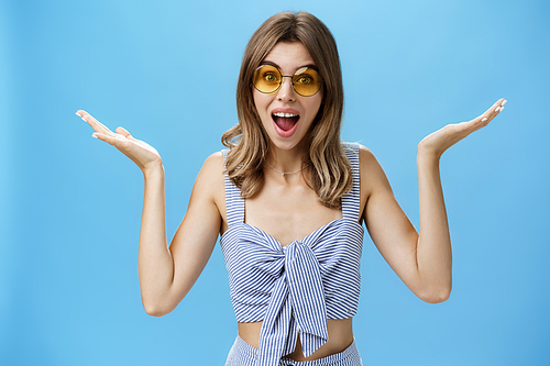 Waist-up shot of charismatic cute young woman in sunglasses raising hands sideways in clueless and unsure gesture shrugging with opened mouth and silly smile being unbothered, posing against blue wall.