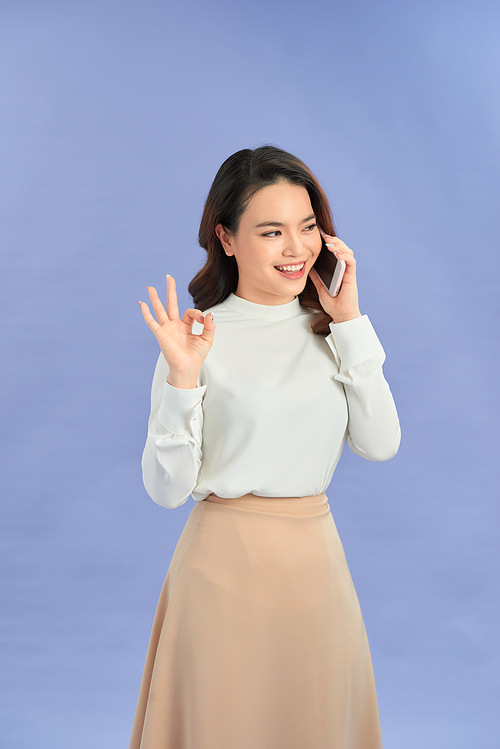 Young woman talking mobile over isolated purple background showing ok sign with fingers