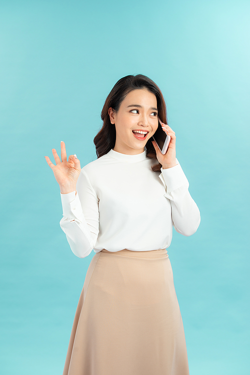 Photo of charming young woman make Okay gesture while talking by her phone over blue background. Look at camera.