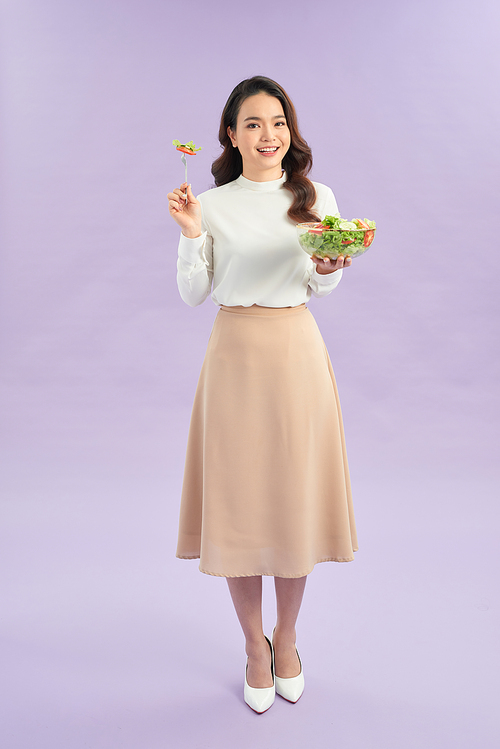 Portrait of a happy playful girl eating fresh salad from a bowl over purple background