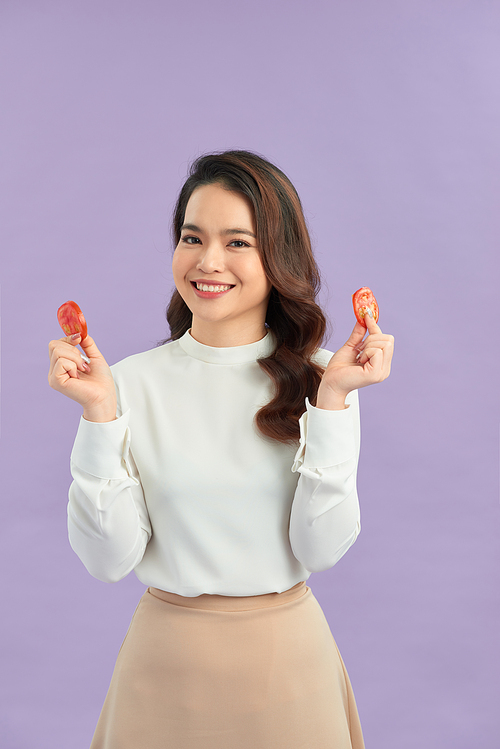 Healthy young Asian woman holding fresh tomato slice over purple isolated background