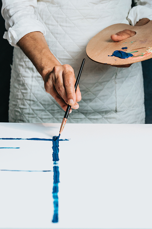 Studio image of an old woman painting a canvas with blue paint. Artist studio concept. Old artist