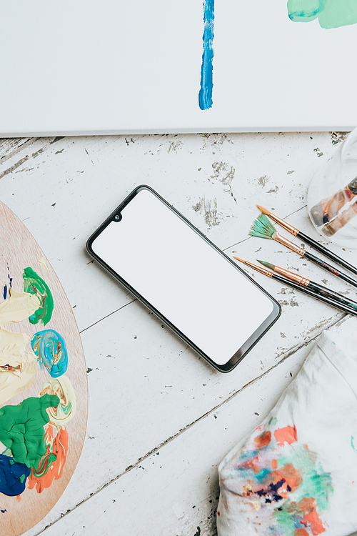 Mock up image of a mobile phone over a white table with a painting palette and painting brushes. Black mobile screen with copy space. Advertising image