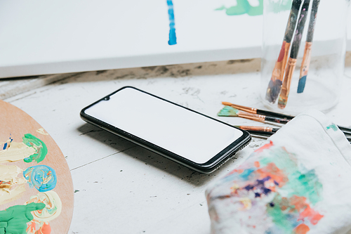 Mock up image of a floating mobile phone over a white table with a painting palette and painting brushes. Black mobile screen with copy space. Advertising image