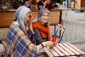 Happy mature woman with grey haired friend rest together sitting at small wooden table in street cafe on nice autumn day