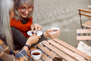 Happy mature woman with glasses takes delicious candy from friend at wooden table in street cafe on nice autumn day