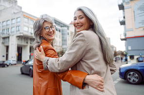 Happy senior Asian woman and grey haired friend hug walking along contemporary city street on autumn day backside view