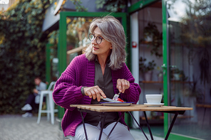 Silver haired mature woman in warm knitted jacket enjoys delicious dessert sitting at table on outdoors cafe terrace on autumn day