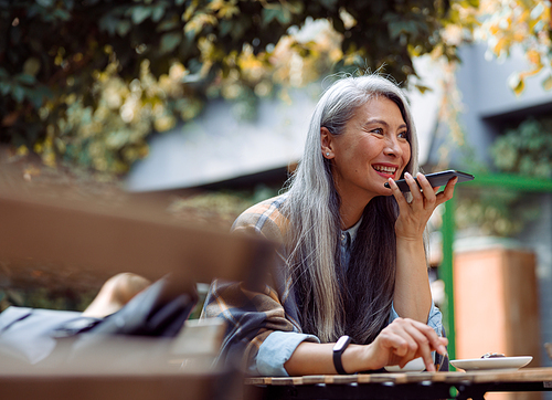 Smiling silver haired Asian lady uses loudspeaker mode on smartphone sitting at table on outdoors cafe terrace on autumn day