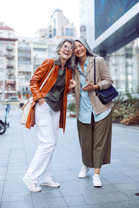 Stylish happy senior women companions stand on modern city street on nice autumn day. Friends spend time together