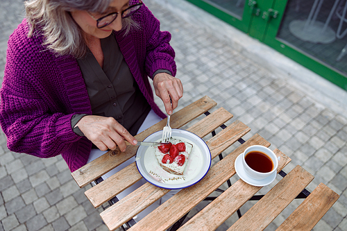 Senior woman in warm purple jacket eats delicious toast with cream and strawberries sitting at table on outdoors cafe terrace on autumn day
