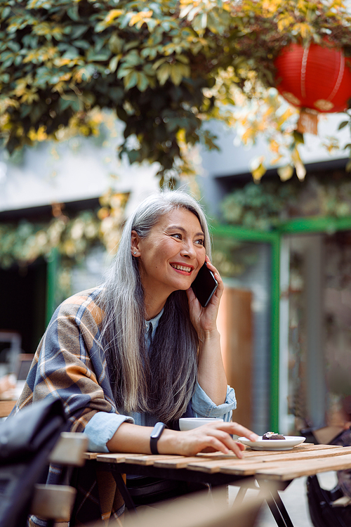 Positive mature Asian woman with hoary hair talks on cellphone resting at small table on outdoors cafe terrace on autumn day