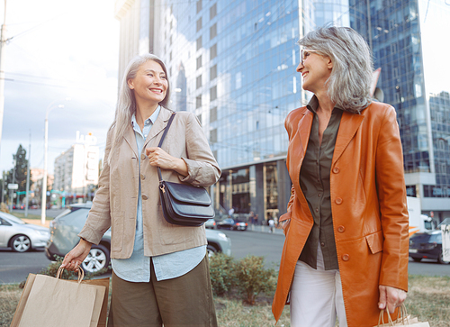 Happy mature ladies in elegant clothes with shopping bags walk along modern city street on autumn day. Friends spend time together