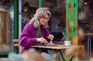 Pretty grey haired senior lady in purple knitted jacket eats delicious dessert sitting at table on outdoors cafe terrace on autumn day