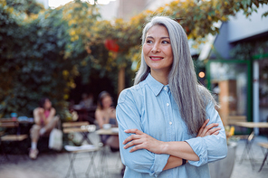Positive hoary haired senior Asian lady with crossed arms in blue blouse stands against outdoors cafe terrace on nice autumn day