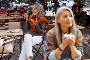 Senior cafe guests, focus on happy silver haired woman with glasses recording audio message on mobile phone on outdoors cafe terrace