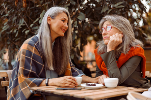 Smiling senior Asian woman holds hand of upset hoary haired friend sitting together at small table in street cafe