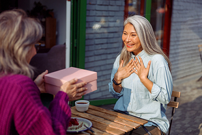 Mature friend gives present to emotional Asian woman sitting at small table on outdoors cafe terrace on autumn day