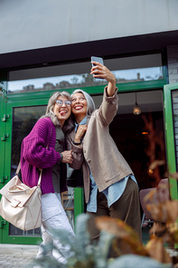 Playful mature woman with Asian friend take selfie showing tongues with mobile phone on modern city street. Long-time friendship relationship