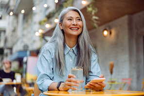 Cheerful mature Asian woman with glass of water and mobile phone sits at small table on outdoors cafe terrace on autumn day