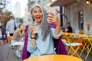 Joyful hoary haired Asian lady with glass of water takes selfie with smartphone at small table on outdoors cafe terrace