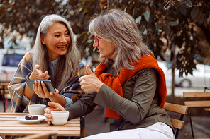 Cheerful senior Asian lady shows phone to grey haired friend at small table with coffee and candies in street cafe on nice autumn day
