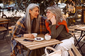 Empathic grey haired Asian woman hugs friend to cheer up sitting together at small table in street cafe on nice autumn day