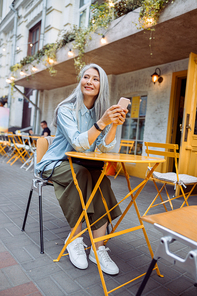 Thoughtful senior Asian woman with silver hair and mobile phone sits at small yellow table on outdoors cafe terrace on autumn day