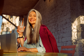 Positive mature Asian woman with grey hair holds drink sitting at small table with open laptop on outdoors cafe terrace at sunset