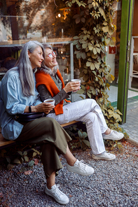 Joyful mature Asian woman with friend hold cups of drinks sitting on bench near cozy cafe window. Long-time friendship
