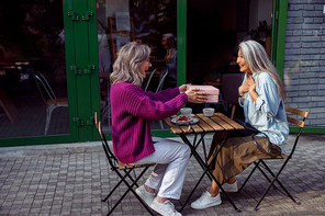 Joyful mature friend gives present to surprised Asian woman sitting at wooden table on outdoors cafe terrace on autumn day