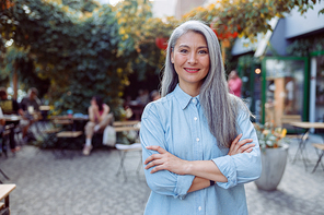 Pretty silver haired senior Asian woman with crossed arms stands against outdoors cafe terrace on nice autumn day
