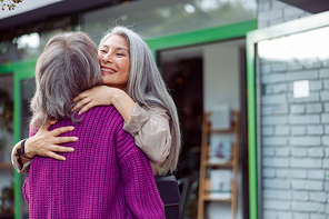 Smiling senior Asian woman hugs female friend in warm purple jacket meeting on modern city street. Long-time friendship relationship