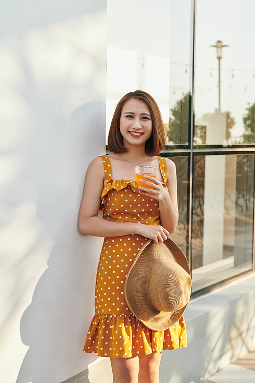 Pretty Asian girl wearing vintage dress and holding hat with orange juice behind wall