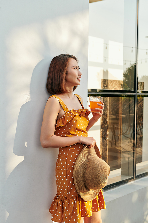 Pretty Asian girl wearing vintage dress and holding hat with orange juice behind wall