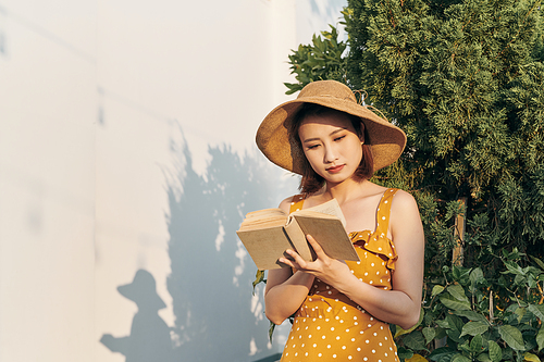 Young woman reading a book standing lean against trunk tree in summer park outdoor.