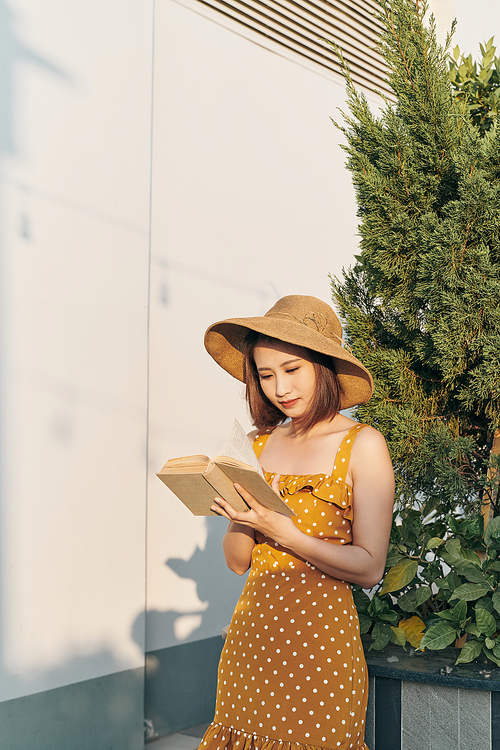 Young woman reading a book standing lean against trunk tree in summer park outdoor.