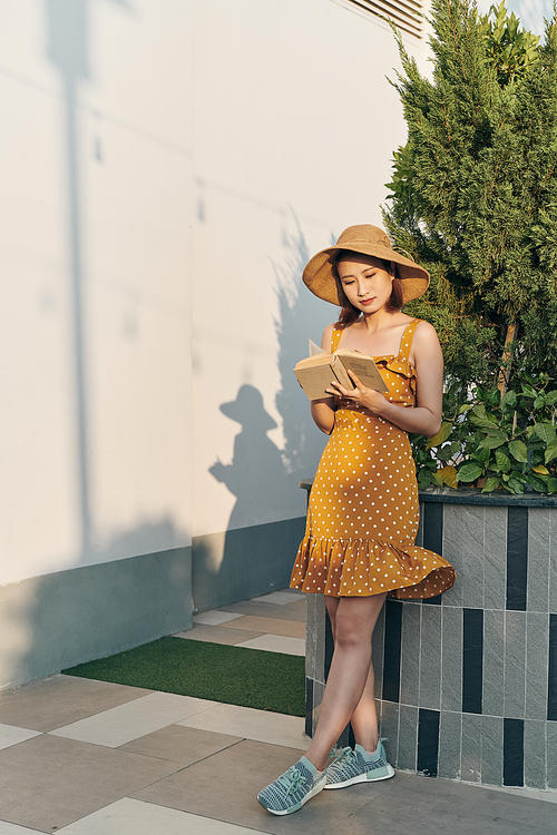 Beautiful young cheerful Asian girl spending good time at the park, reading paper book while standing