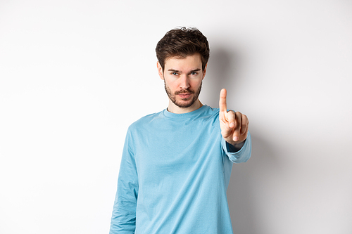 Image of serious young man with beard, shaking finger in disapproval, prohibit or forbid something, standing over white background and saying no.