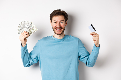Happy guy smiling and showing plastic credit card with cash, choosing between money and contactless payments, white background.