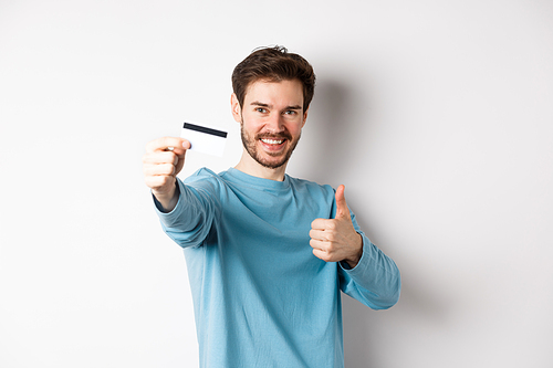 Handsome caucasian man showing plastic credit card with thumb-up, recommend and praise good bank offer, standing on white background
